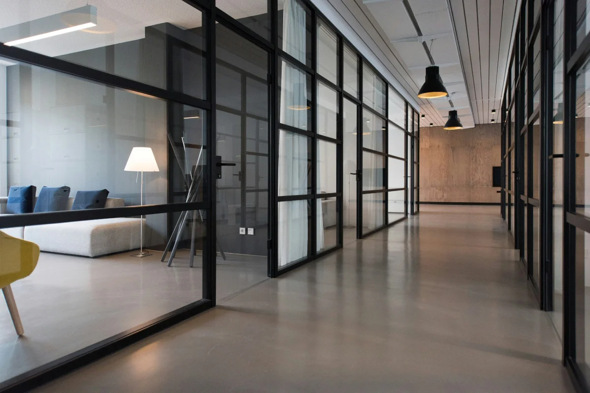 Long glass-walled office corridor with black metal frames and a seating area to the left, including a white sofa and floor lamp.
