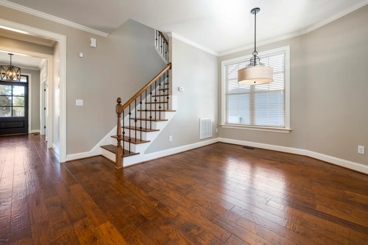 Bright empty living room with hardwood floors, a staircase with wooden railing, and a large window with blinds; contemporary chandelier hangs from the ceiling.