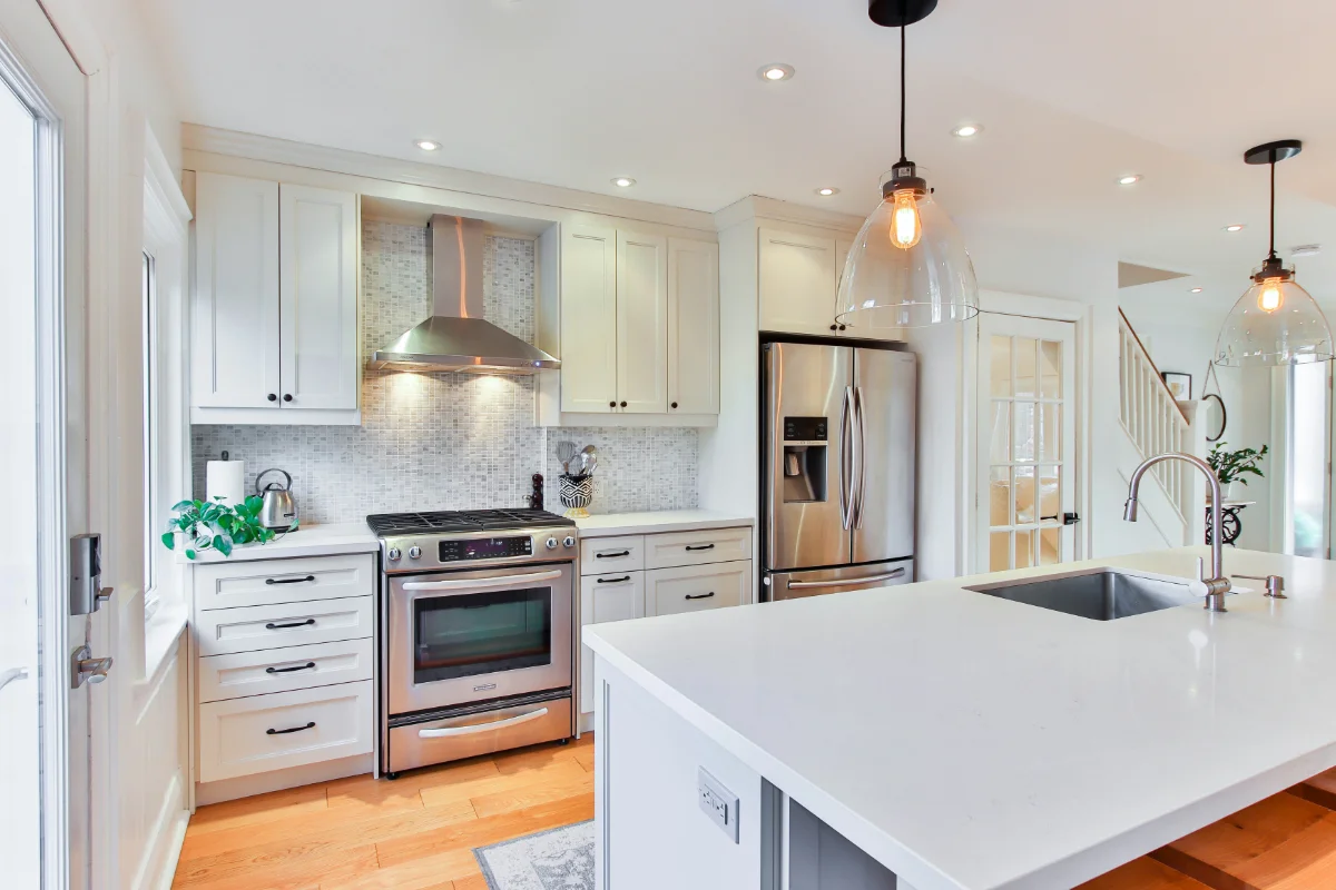 Bright modern kitchen with white cabinets, stainless steel fridge and range, and a large white island with a sink and pendant lights above.